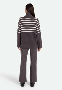 Pull en tricot à rayures gris foncé avec col haut, présentant de larges rayures blanches. Associé à un pantalon gris évasé et des chaussures noires à semelles épaisses.