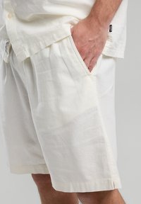 Pantaloni corti bianchi in lino con una vestibilità rilassata, tasche laterali e orlo piegato. Il tessuto ha una texture liscia con lievi pieghe.