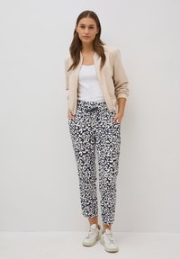 Giacca bomber beige chiaro sopra una canotta bianca, abbinata a pantaloni blu navy con un motivo astratto bianco e sneakers bianche.