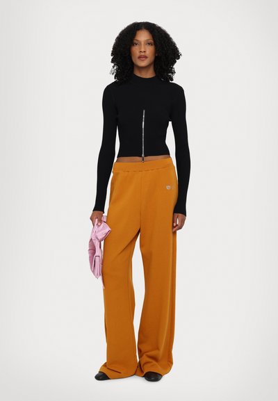 Fekete bordás crop top elülső cipzárral, széles narancssárga nadrággal párosítva. A modell egyik kezében egy rózsaszín tárgyat tart. Az anyagok sima textúrájúak.