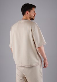 Beige oversized short-sleeve shirt with a round neckline, featuring textured fabric and contrasting stitched hems. Soft, casual design.