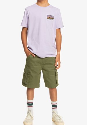 Jasnofioletowy bawełniany T-shirt z małym graficznym wzorem na kieszeni, zestawiony z oliwkowo-zielonymi szortami cargo z wieloma kieszeniami. Białe skarpetki w paski.