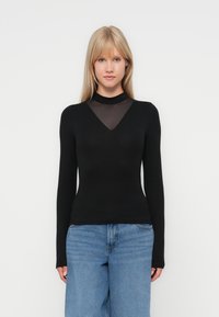 Black long-sleeve top with a V-neckline, featuring a sheer mesh panel at the neck and fitted silhouette. Paired with light blue denim.