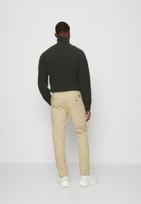 Dunkelgrüner, gerippter Pullover mit hohem Kragen, kombiniert mit beigen Chinos und weißen Sneakern. Keine auffälligen Muster oder Akzente sichtbar.