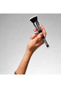 Pennello per il trucco con una ghiera in metallo argento, manico nero e setole morbide con una sfumatura dal viola al bianco, tenuto in mano.