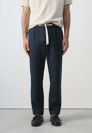 Hombre con pantalones azul marino con cordón, zapatos negros y una camisa beige de manga corta, de pie frente a un fondo liso.