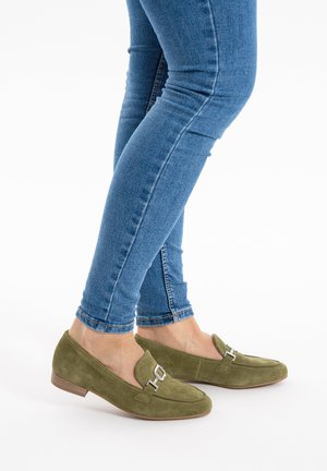 Person wearing blue skinny jeans and olive green suede loafers with silver buckle details, standing on a white background.