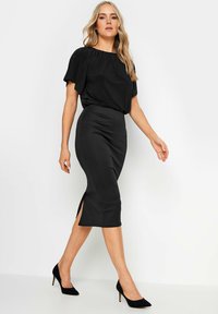 Robe noire avec des manches chauve-souris et une jupe crayon cintrée avec une fente sur le côté. Texture lisse et longueur midi, associée à des talons noirs.