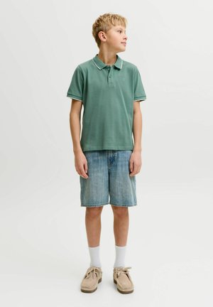 Jongen met blond haar die een groen poloshirt, spijkershorts, witte sokken en beige schoenen draagt, staat en kijkt opzij.