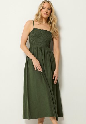 Lyshåret kvinde iført en ærmeløs maxi-kjole i olivengrøn med blomsterbroderi på overdelen, stående mod en enkel lys væg.