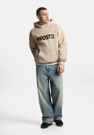 Młody mężczyzna w beżowej bluzie z kapturem „PROSTO.”, jasnoniebieskich szerokich dżinsach i czarnych trampkach, stojący na jednolitym białym tle, patrzący na bok.