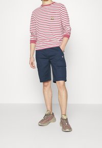 Muž v růžovém a bílém pruhovaném tričku s dlouhým rukávem, námořnických modrých cargo šortkách a béžových a růžových teniskách, stojící na bílém pozadí.