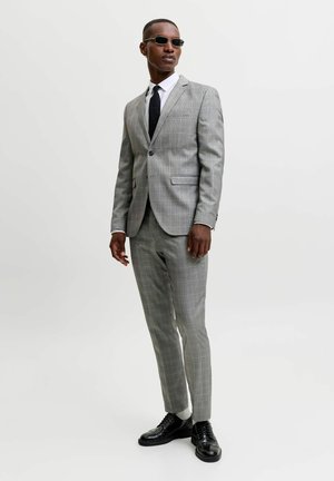 Homme portant un costume gris à carreaux, une chemise blanche, une cravate noire, des chaussures noires, des chaussettes blanches et des lunettes de soleil foncées, debout devant un fond uni.