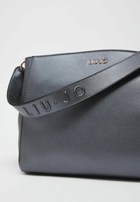 Sac à main en cuir noir texturé avec un bord cousu, doté d'une large sangle embossée et d'un détail de logo doré. Design compact et angulaire.