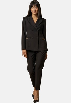 Un blazer doppiopetto nero su misura presenta dettagli in hardware dorato e due tasche con zip, abbinato a pantaloni neri aderenti e tacchi.