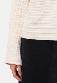 Un pull crème à mailles torsadées, avec des manches et un torse, associé à une jupe noire, montrant une main avec des ongles vernis nude sur le côté.