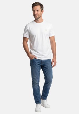 Mand står med hånden i lommen iført hvid T-shirt med korte ærmer, blå jeans og hvide sneakers på en hvid baggrund.
