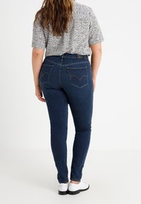 Jeans de un azul oscuro con un corte ajustado, que cuentan con bolsillos traseros y un sutil detalle de costura. Llevados con una camisa de patrón en blanco y negro.