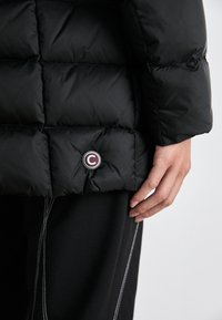 Zwarte pufferjack met een geborduurd ontwerp, met een ronde logopatch nabij de zoom. De stof heeft een gladde textuur en is lichtgewicht.