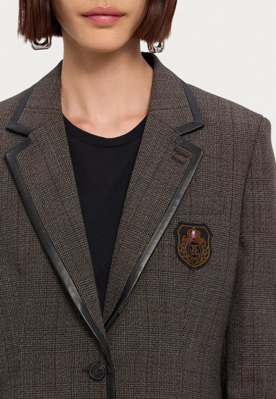 Femme portant un blazer marron à carreaux avec bordure en cuir noir et patch écusson brodé, sur une chemise noire, montrant le cou et le bas du visage.
