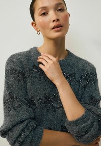 Szary, fakturowany sweter z miękkiego, puszystego materiału, z czarnymi cekinami ułożonymi w wzór na przodzie i rękawach.