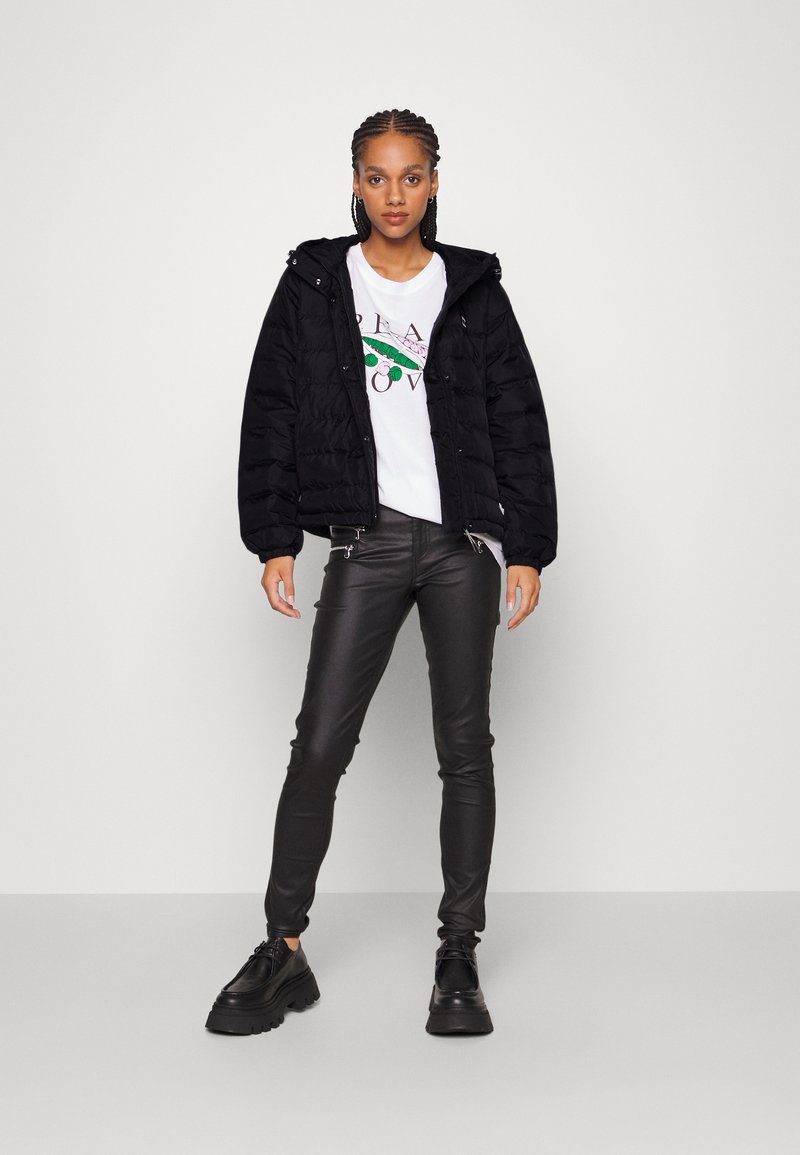 Doudoune noire avec capuche, texture matelassée et fermeture éclair sur le devant. Portée par-dessus un t-shirt graphique blanc et associée à un pantalon en faux cuir noir et des chaussures.