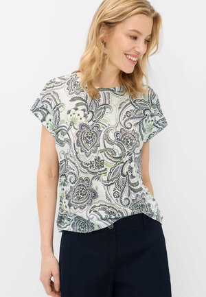 Donna bionda che sorride, indossa una maglietta bianca a maniche corte con motivi paisley verdi e neri e pantaloni blu scuro.