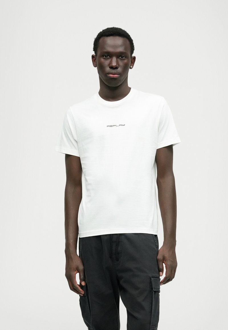 Biała koszulka z krótkim rękawem z małym czarnym logo "REPLAY", noszona z czarnymi spodniami cargo, prezentująca gładką teksturę i klasyczny krój.