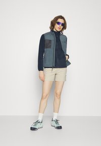 Giacca leggera con pannelli blu e neri sopra un pile blu navy, abbinata a pantaloni corti beige e scarpe da trekking fucsia. Occhiali da sole specchiati completano il look.