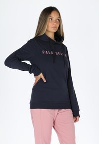 Felpa della marina con la scritta "PALM BEACH" in stampa rosa, cappuccio con cordini, texture morbida e polsini a coste, abbinata a pantaloni rosa chiaro.