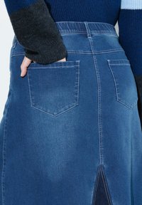 Falda de mezclilla en azul medio con cintura ajustada, bolsillos en la parte trasera y un sutil detalle de abertura en el dobladillo. Textura suave.
