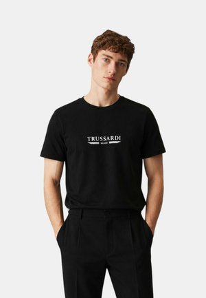 Giovane uomo con capelli ricci che indossa una t-shirt nera Trussardi Milano e pantaloni neri, in piedi con le mani nelle tasche su uno sfondo bianco.