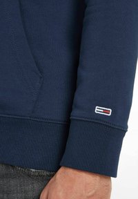 Felpa blu navy con polsini a costine, caratterizzata da un piccolo logo ricamato con accenti rossi, bianchi e blu vicino al polso.