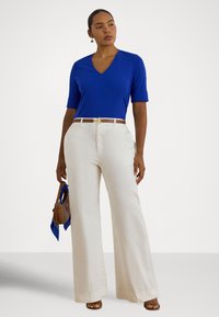 Žena v modrom tričku s výstrihom do V a rukávmi po lakte, s vysokým pásom a širokými bežovými nohavicami, drží hnedú kabelku s modrým šálom.