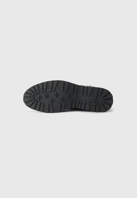 Black rubber shoe sole with a textured grip pattern, featuring deep treads and raised sections for traction. Clean white background.