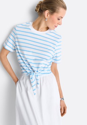 Vrouw met blond haar in een knot, draagt een blauw-wit gestreepte top met knoop, witte rok, zilveren oorbellen en een zwart-witte armband, kijkt omlaag.