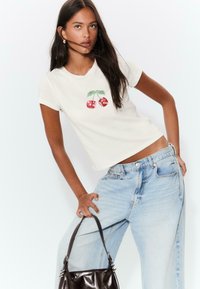 T-shirt bianco corto con grafica di ciliegia rossa, maniche corte e scollatura rotonda. Abbinato a jeans chiari a vita alta e una borsa scura.