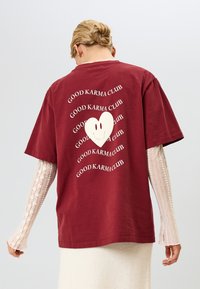 Bordo oversize majica s velikim belim srcem sa smeškom i ponovljenim tekstom "DOBRA KARMA KLUB" u talasastom uzorku. Pamuk.