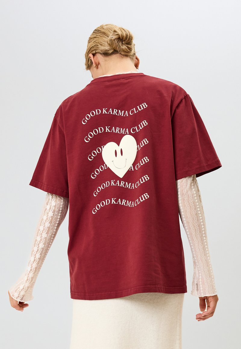 Bordo oversize majica s velikim belim srcem sa smeškom i ponovljenim tekstom "DOBRA KARMA KLUB" u talasastom uzorku. Pamuk.