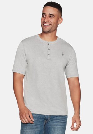 Lächelnder Mann, der ein hellgraues kurzärmeliges Henley-Shirt mit drei Knöpfen und blaue Jeans vor einem schlichten Hintergrund trägt.