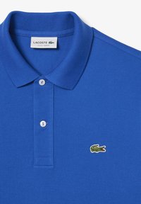 Blå polo-r skjorta i texturerad tyg, med en klassisk krage, två knappar och en grön krokodil-logotyp på vänster bröst.