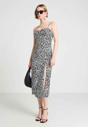 Donna che indossa un vestito midi nero e bianco a pois con spacco laterale, tacchi neri, occhiali da sole, orecchini floreali bianchi e tiene una borsa nera.