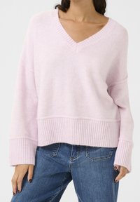 Pull en tricot rose clair avec un décolleté en V, des poignets et un ourlet côtelés. Présente une coupe décontractée et une texture douce, associé à un jean en denim bleu.