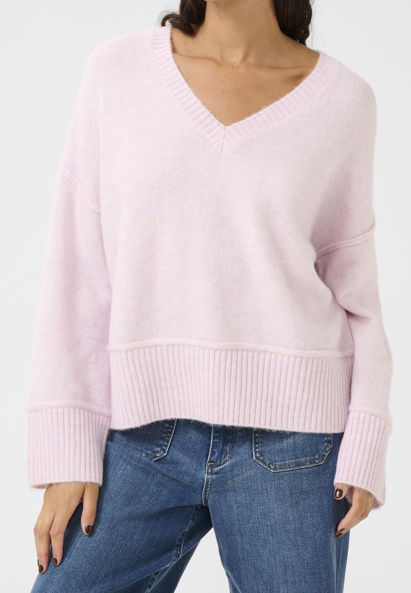 Pull en tricot rose clair avec un décolleté en V, des poignets et un ourlet côtelés. Présente une coupe décontractée et une texture douce, associé à un jean en denim bleu.