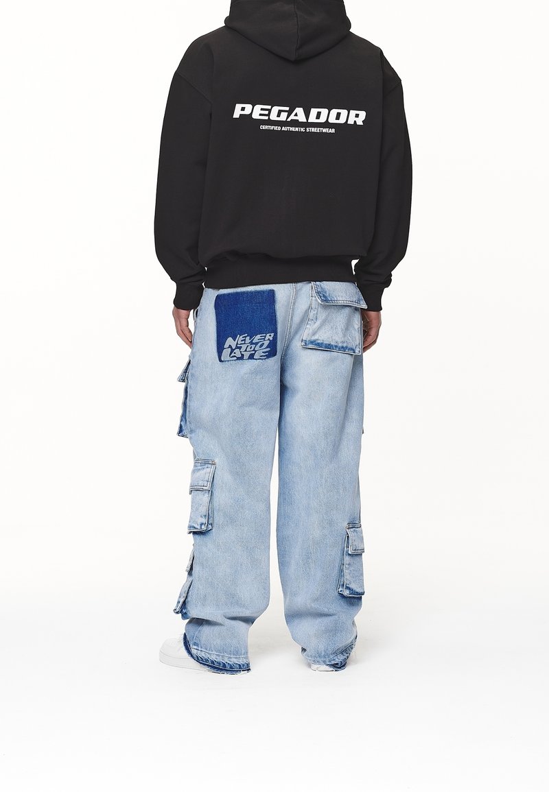 Musta huppari, jossa on "PEGADOR" -logo selässä, yhdistetty vaaleansinisiin cargo-farkkuihin, joissa on kirjontatyylinen koriste ja tekstit oikeassa takataskussa.