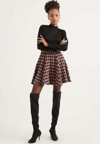 Top nero a maniche lunghe abbinato a una gonna svasata a quadri rosa e neri. La modella indossa stivali neri con tacco sopra il ginocchio, con le braccia incrociate.