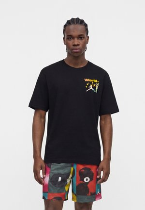 Mužský model s vrkočmi nosí čierne tričko s farebným logom "World Tour" a živé viacfarebné vzorované šortky, stojaci na bielom pozadí.