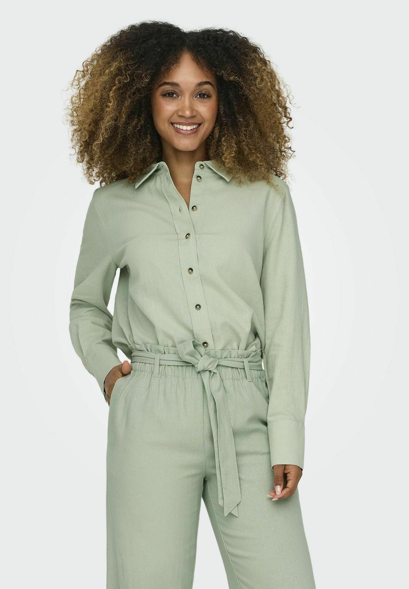 Camicia verde chiaro, botonata e con colletto, abbinata a pantaloni a vita alta coordinati con cintura e tasche laterali. Texture morbida.