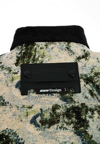 Chaqueta de tela texturizada en beige con patrones abstractos en verde y azul. Presenta un collar de pana negro y una etiqueta de tela negra con la marca.