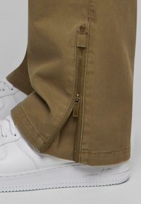 Beige bomullsmixbyxor med dragkedja vid vristerna, i kombination med vita sneakers. Har en rak passform och en mjuk textur.